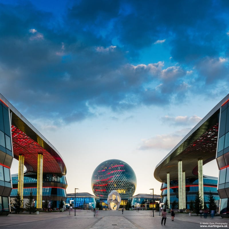 Veža Bajterek v Astane nasvietená v noci s modernou panorámou mesta.
