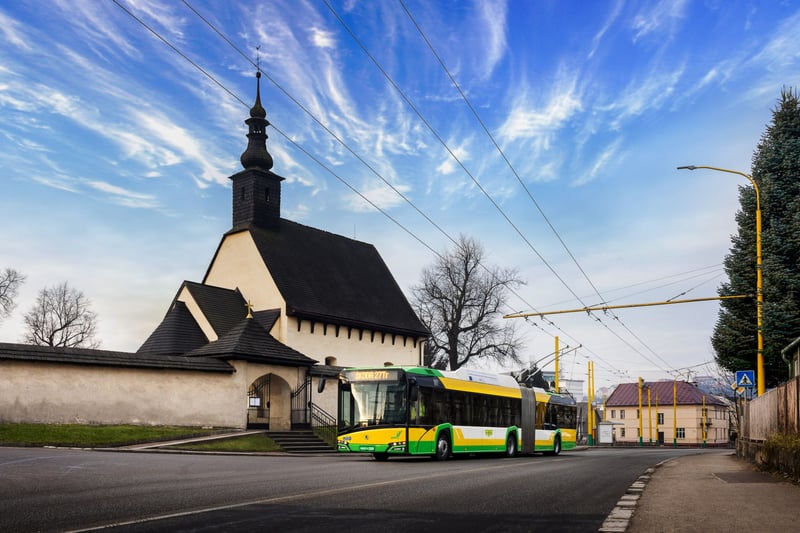Žltozelený trolejbus Solaris MHD Žilina zaparkovaný pri vidieckej kaplnke v lete.