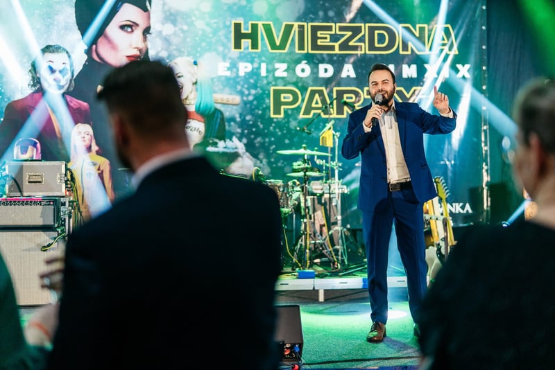 Spevák v modrom saku s mikrofónom na pódiu tematickej firemnej party, v popredí publikum