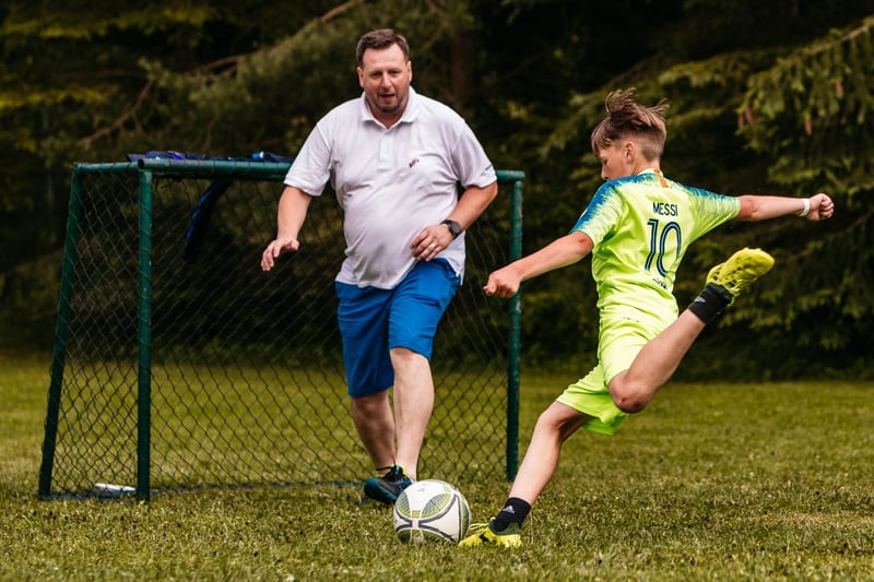 Otec a jeho syn v zelenom futbalovom drese strieľajú gól pri bránke počas family day v prírode