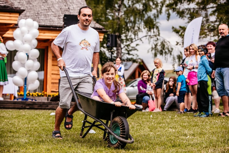 Otec tlačí dcéru v záhradnom fúriku počas family day-u, v pozadí biele balóny a chata