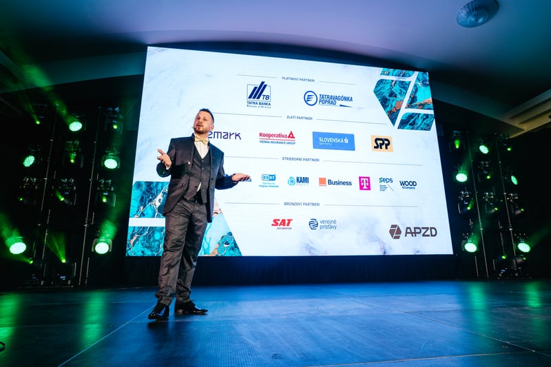 Moderátor v obleku gestikuluje na pódiu pred slide so zoznamom partnerov a sponzorov gala večera