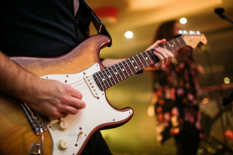 Detail rúk gitaristu hrajúceho na elektrickej gitare, v pozadí kapela