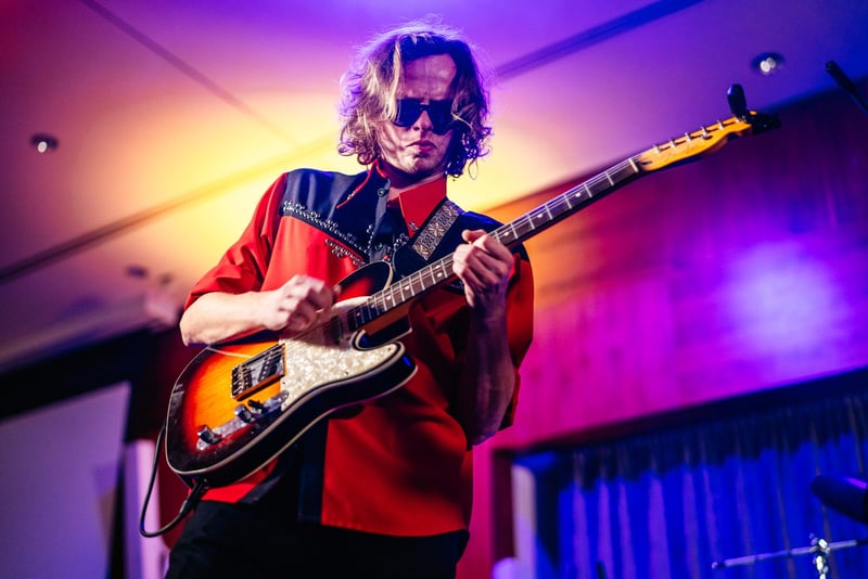 Gitarista s dlhými vlasmi a slnečnými okuliarmi v červenom kostýme hrá na elektrickej gitare v purpurovom svetle