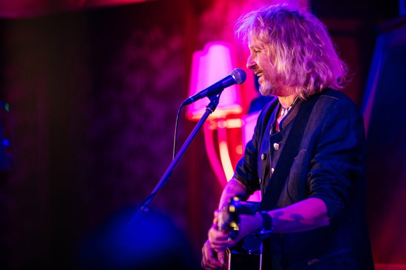 Spevák s dlhými vlasmi a akustickou gitarou pri mikrofóne vo fialovom osvetlení