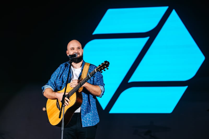 Spevák s akustickou gitarou v košeli pri mikrofóne na pódiu pred LED brand panelom slávnostného večera