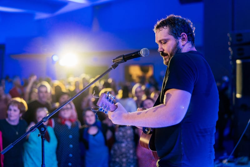 Spevák s akustickou gitarou na pódiu pred tlieskajúcim publikom