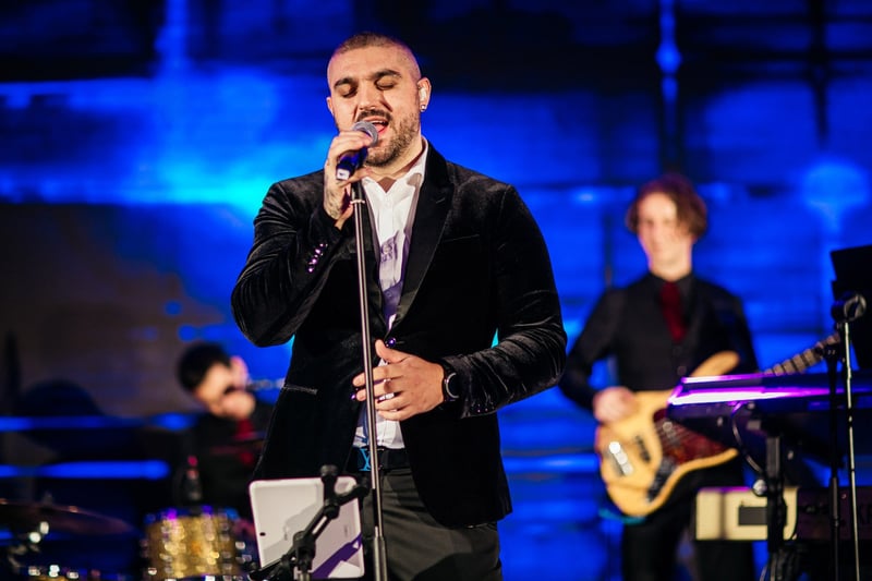 Spevák v čiernom saku spieva pri mikrofónovom stojane, v pozadí basgitarista a kapela