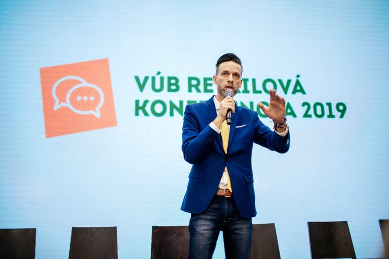 Rečník v modrom saku s mikrofónom v ruke gestikuluje pred slide-om konferencie
