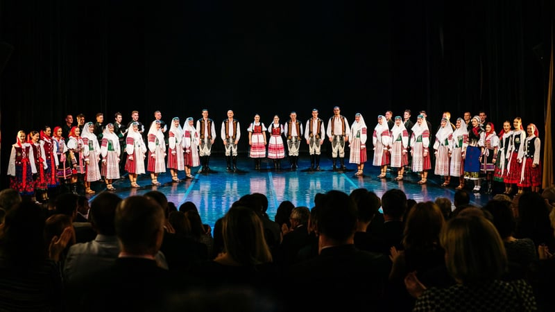 Celý folklórny súbor v krojoch stojí v rade na divadelnom pódiu pred sediacim publikom