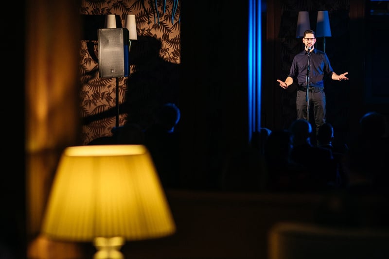 Speaker v tmavom outfite s mikrofónom v intímnom hotelovom salóne pred publikom v tieni
