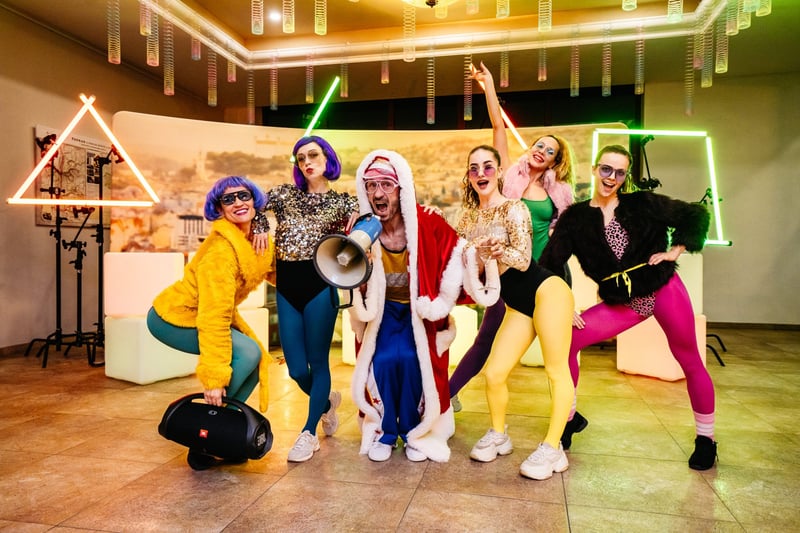 Skupina v 80s kostýmoch s parochňami a neónovými outfitmi pózuje s mikrofónom na tematickej firemnej party